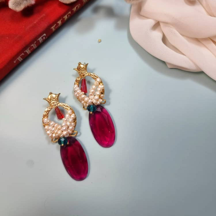 A pair of gold-tone drop earrings with white faux pearls, a small red gem, and a large oval magenta pink drop, displayed on a light blue surface with decorative props.