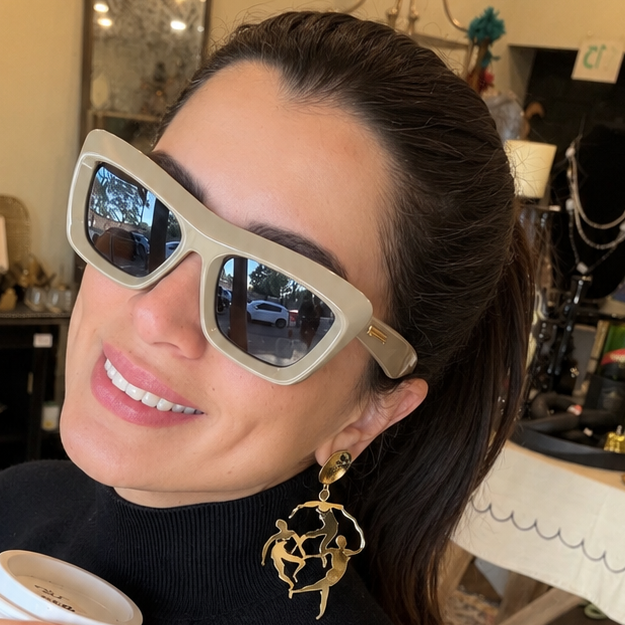 A woman wearing chunky off-white square sunglasses and a black turtleneck sweater, holding a white coffee cup in a cozy, cluttered indoor setting.