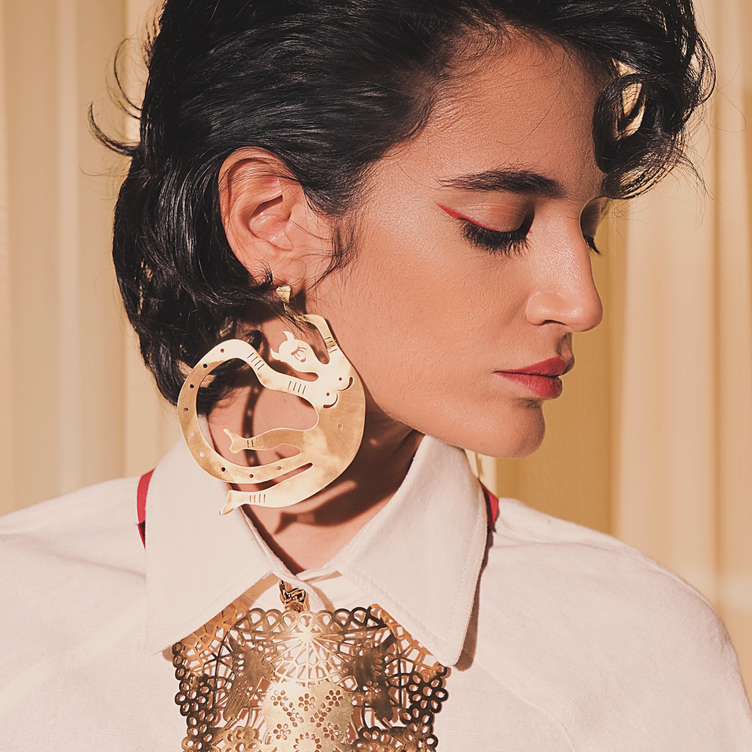 A close-up portrait of a model wearing dramatic, large gold-toned statement earrings and a matching ornate gold collar necklace with red bead fringe, paired with a white collared shirt.
