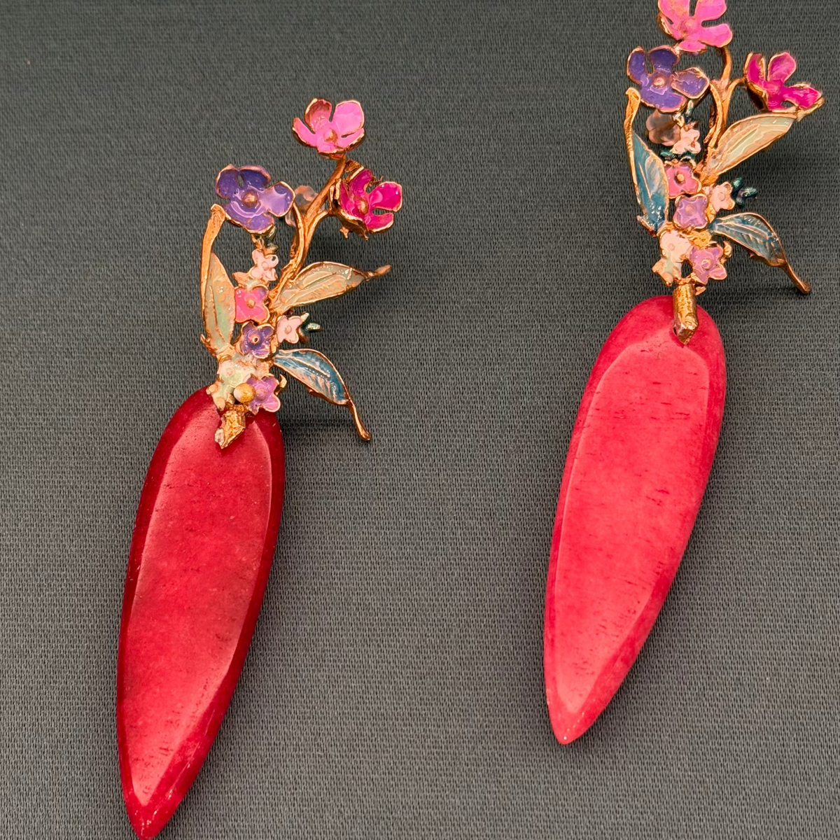 A pair of red teardrop-shaped earrings with gold-toned floral details and small pink and purple flowers, displayed on a dark gray textured background.