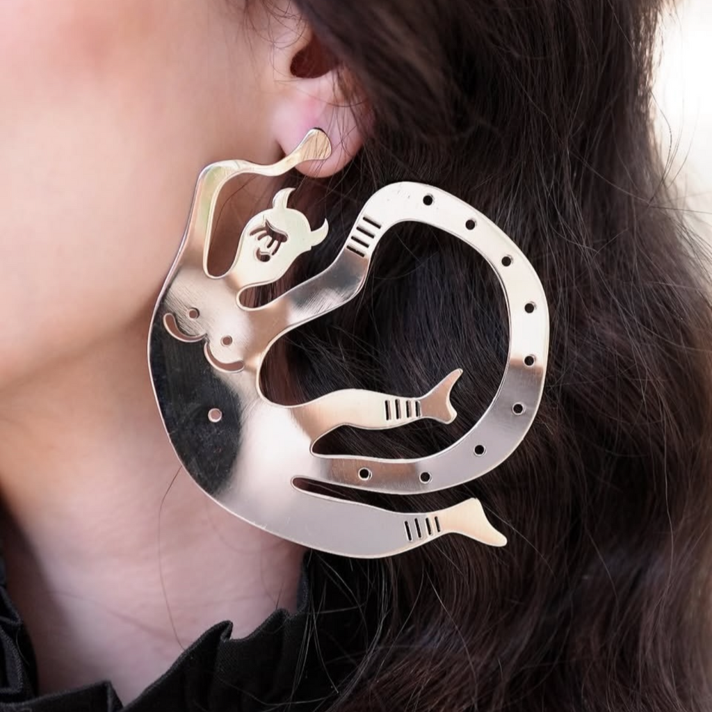 A close-up of a person wearing a large, unique metallic earrings, The earring has a silver-toned finished with delicate perforated details,