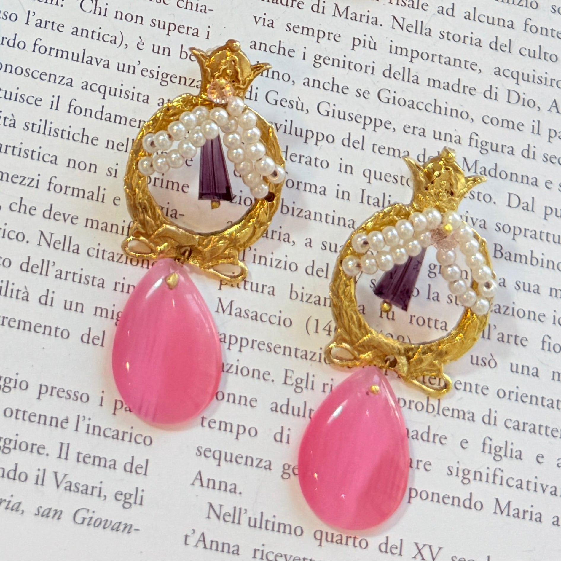 A styled flat-lay of six ornate gold-tone earrings with colorful gemstone and bead details, arranged on an open book with Italian text. The earrings feature purple, red, black, and pink stone drops with gold filigree and crown-like settings.