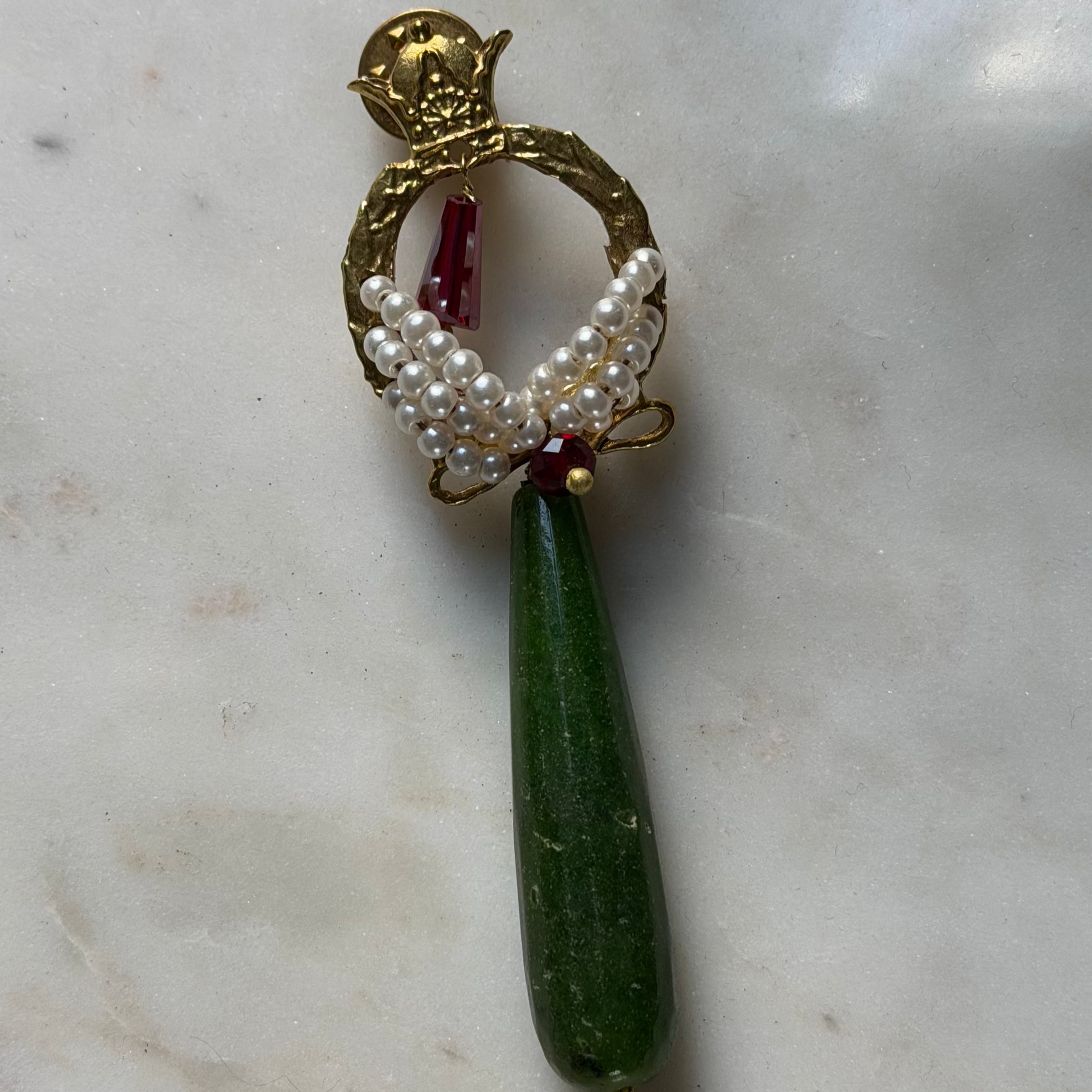 A vintage gold-tone brooch featuring a dangling green teardrop-shaped stone, a pearl-encrusted wing design, and a small red gemstone accent, displayed on a light marble surface.