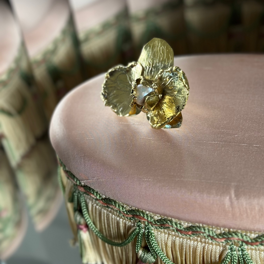 A BRASS ring shaped like a ORCHID with a central PEARL, resting on the pink velvet top of an upholstered stool.