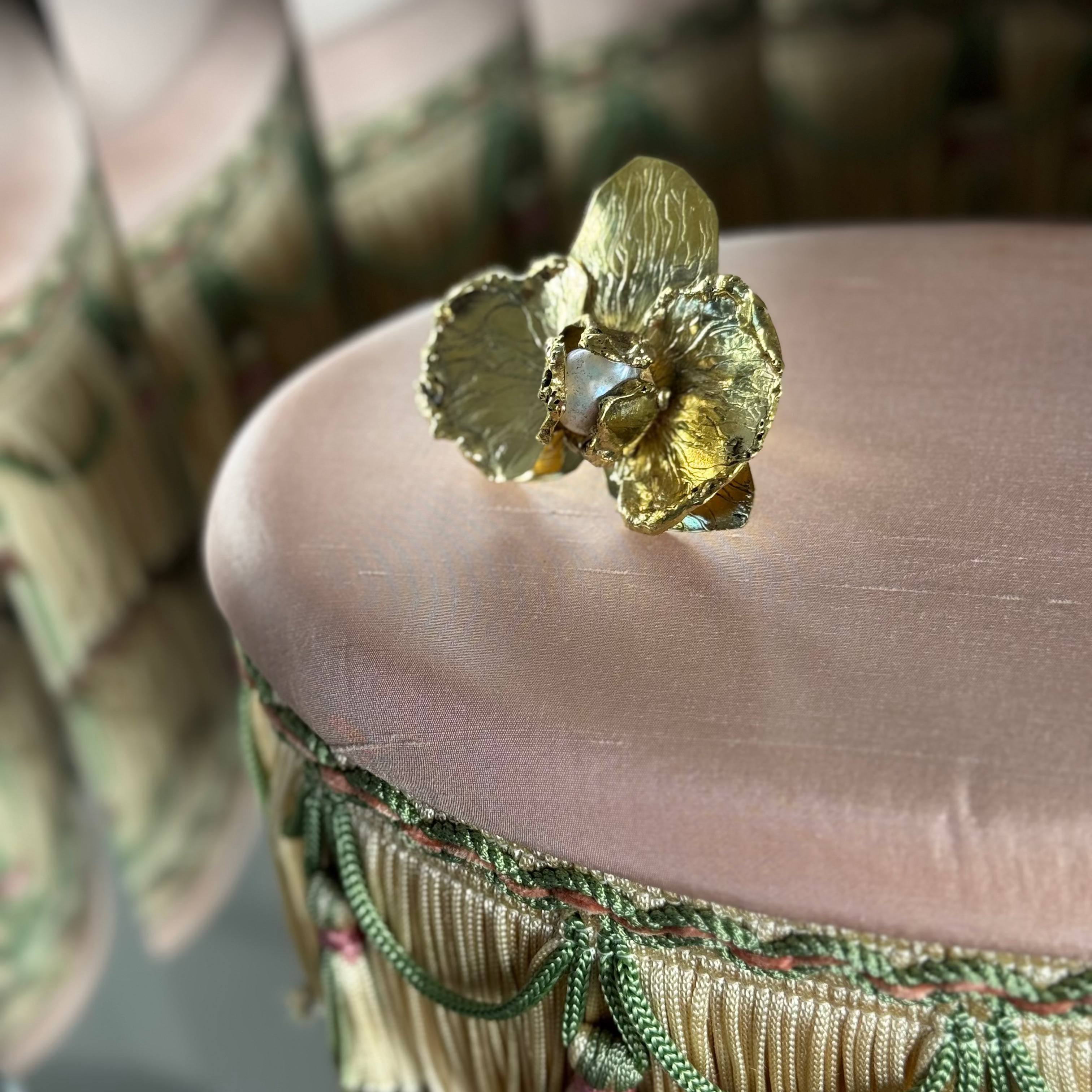 A BRASS ring shaped like a ORCHID with a central PEARL, resting on the pink velvet top of an upholstered stool.
